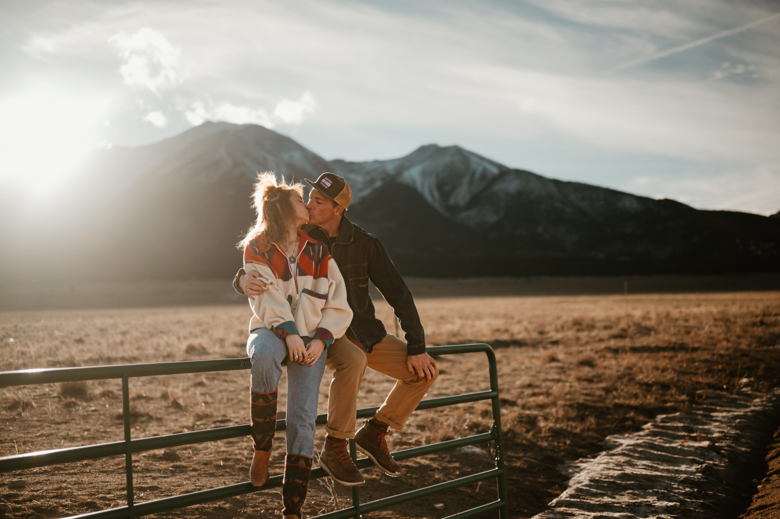 Colorado Mountains with a beautiful couple sitting on a fence kissing with soft warm sunlight on them